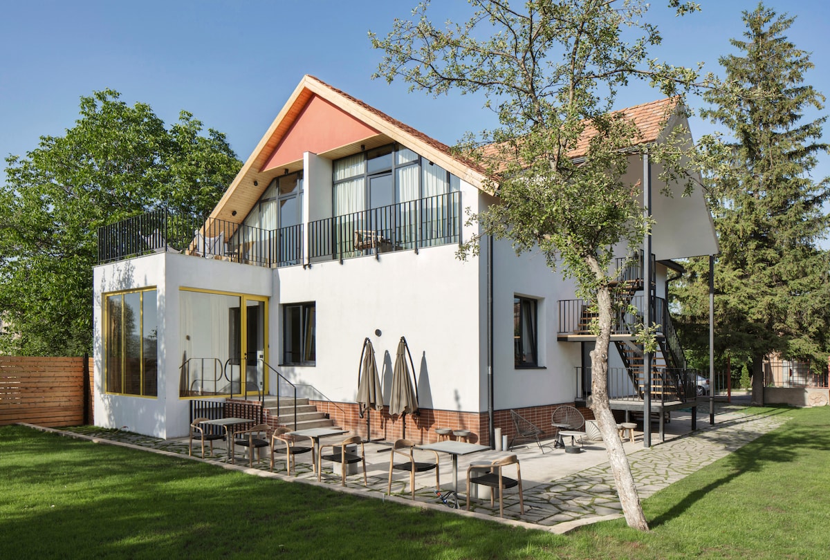 A modern two-story house features a combination of white and red exterior with large windows. A patio area is adorned with several lounge chairs and umbrellas, surrounded by well-maintained grass and trees. A staircase leads to a second-floor balcony with railings.