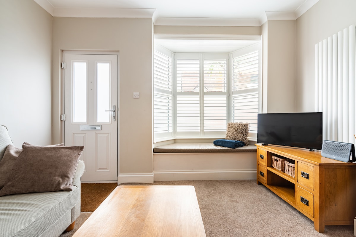 A bright living area features a comfortable sofa and a large window with wooden shutters, allowing natural light to filter in. A wooden coffee table is positioned in front, while a TV is set on a stylish cabinet. A cozy cushion rests on the window seat.