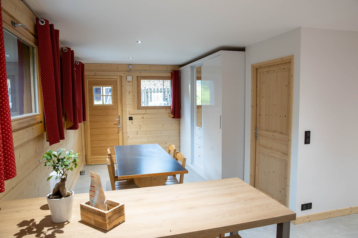 A well-lit dining area features a large wooden table surrounded by light wooden chairs. The space is enhanced by warm wooden walls and red curtains, with a door leading to the outside. A small plant and a wooden storage box add a touch of warmth.