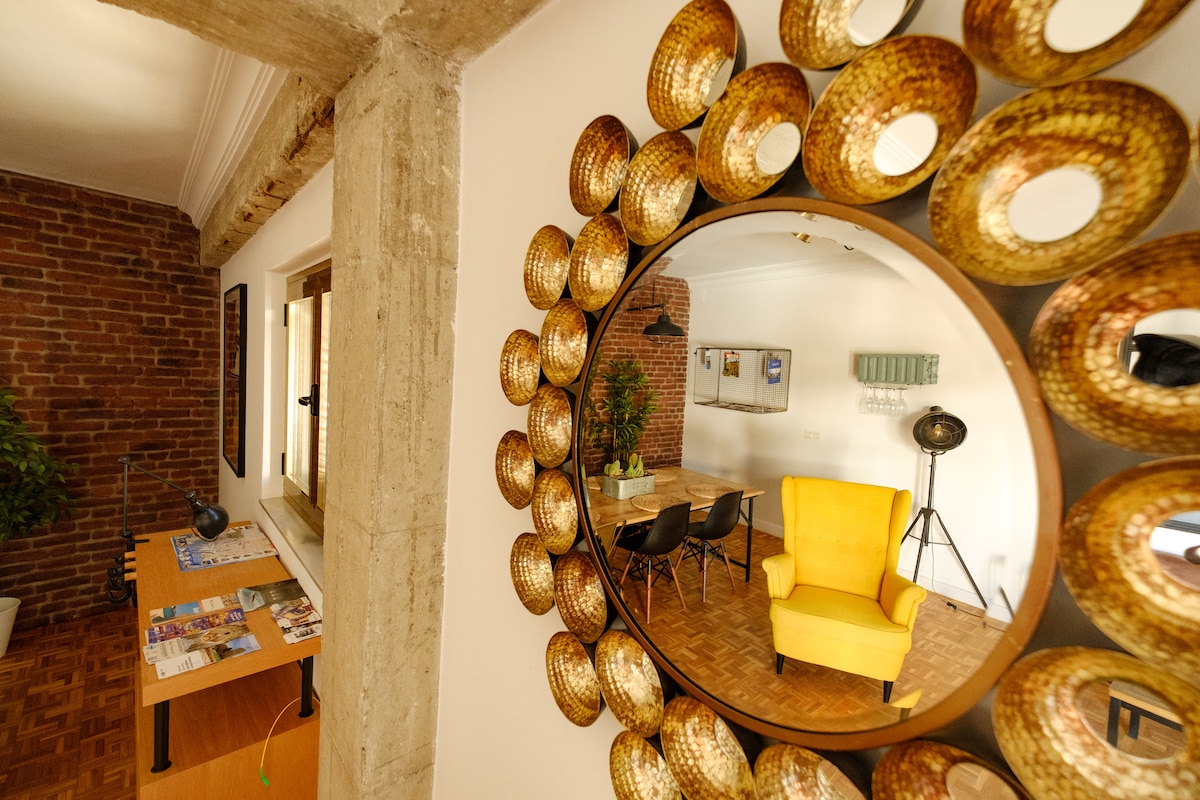 A round mirror is framed by metallic circles, reflecting a modern arrangement. A bright yellow chair adds a pop of color beside a wooden table set for meals. Brick walls provide an industrial touch, while a small plant brings a hint of greenery to the space.