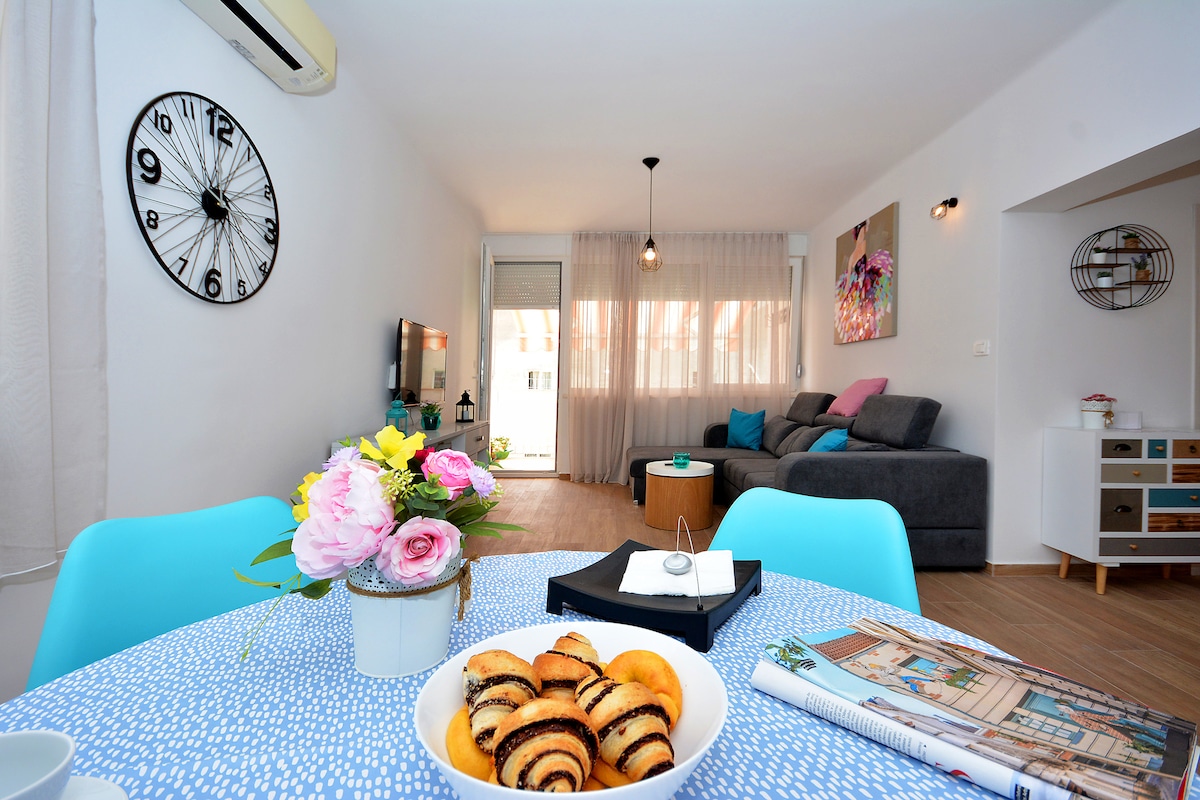 A modern dining area features a round table set with a white bowl of pastries, surrounded by vibrant blue chairs. The living space beyond showcases a gray sofa, a decorative wall clock, and a colorful wall art piece. Natural light filters through sheer curtains.