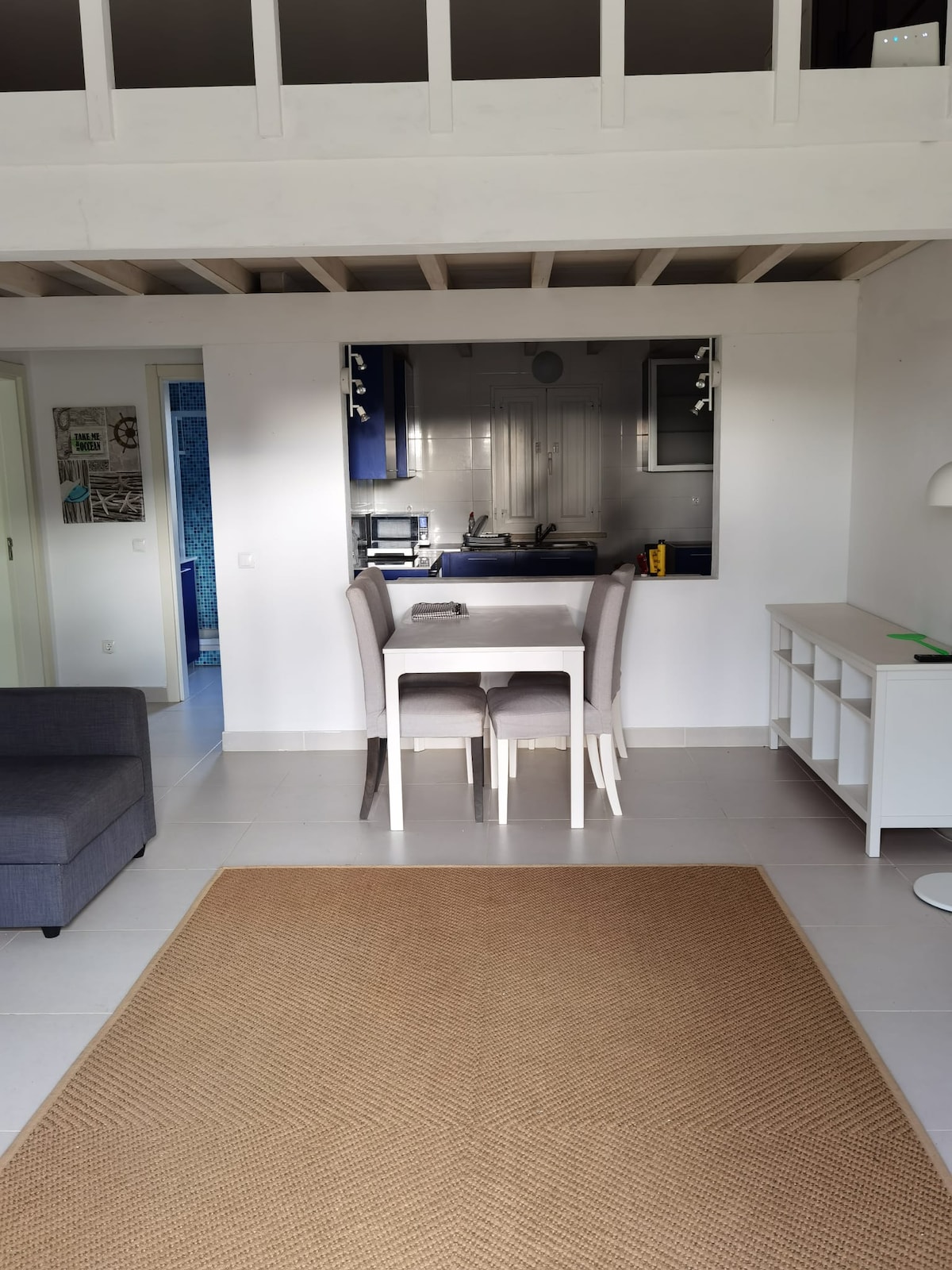 A dining area is presented with a white table surrounded by four chairs, positioned across from a kitchenette with modern appliances. A gray couch and a light-colored rug enhance the spaciousness of the room, which features white walls and wooden beams above.