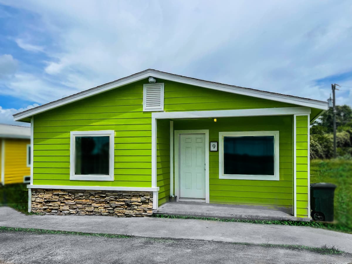 The exterior of a cottage is presented, featuring a vibrant green façade with white trim and a stone accent at the base. Two large windows flank the entrance, providing ample light inside. A concrete pathway leads to the front door, complemented by a grassy area beside the driveway.