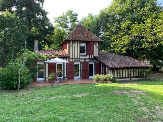 The exterior features a charming, traditional structure with exposed wooden beams and a tiled roof. Lush greenery surrounds the house, with a spacious terrace adorned with white curtains. The inviting garden space is punctuated by vibrant plants, offering a serene setting amidst nature.