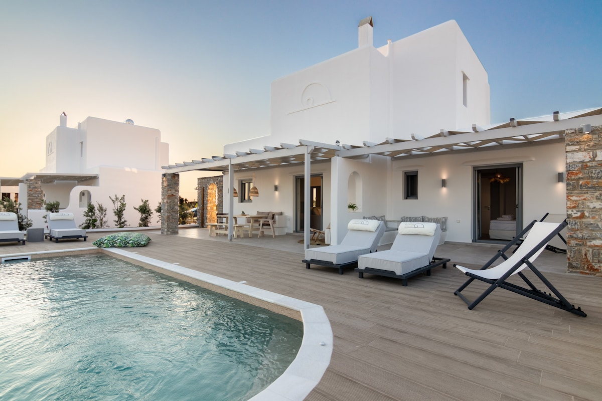 The exterior of a modern two-story villa is showcased, featuring a private swimming pool surrounded by sunbeds. A shaded outdoor patio with a dining table is visible beneath a wooden pergola. The villa's whitewashed walls reflect the sunlight amid a backdrop of clear skies.