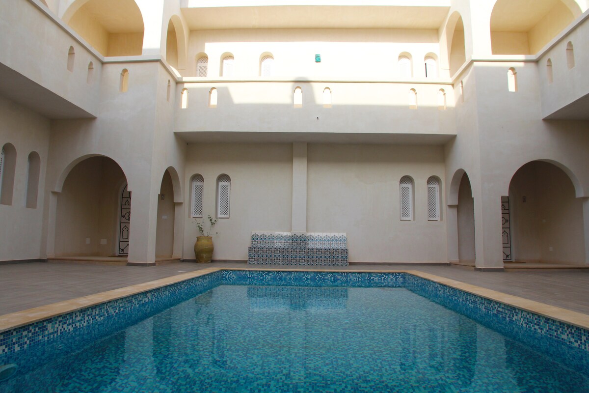 An indoor pool area is highlighted by smooth, shimmering blue water. Surrounding arches and light-colored walls create an airy ambiance, while decorative tiles are positioned along one side. Comfortable seating is available in the background, providing a place to relax by the water.