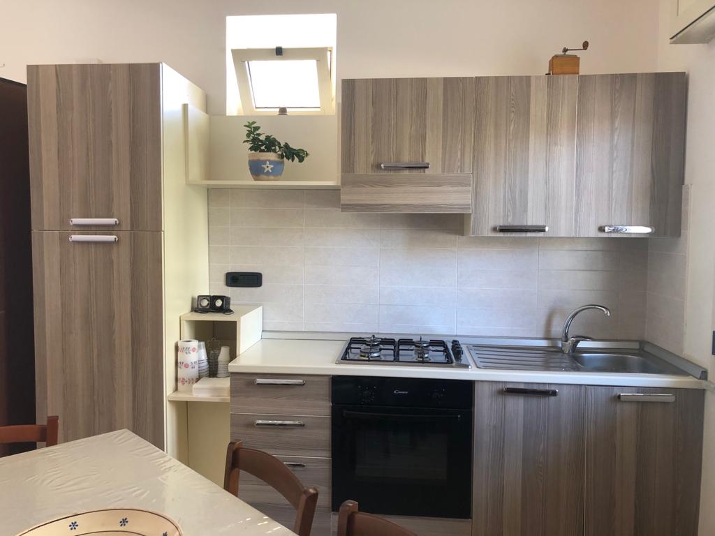 The kitchen features modern cabinetry in natural wood tones, with a gas stove, oven, and sink integrated into the countertop. A small window above the sink lets in natural light, while a potted plant adds a touch of greenery.