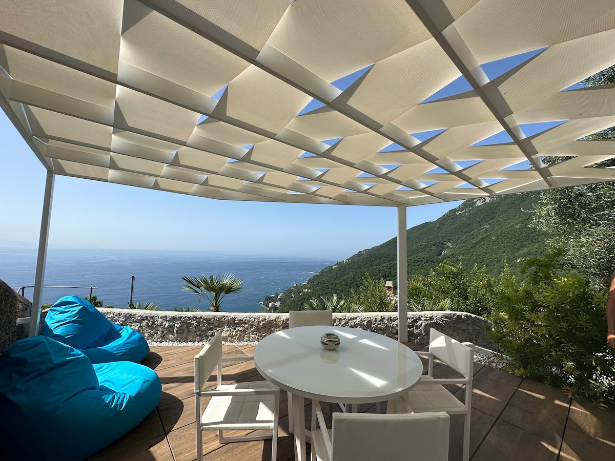 An outdoor dining area is set under a modern canopy, providing shade while offering views of the sea and surrounding hills. A round table is surrounded by white chairs, with two blue bean bags placed nearby for relaxation.