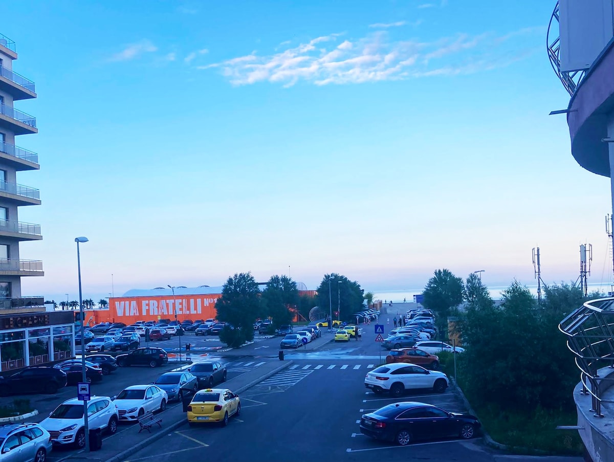 A view overlooking a parking lot with various vehicles parked. The road leading to the beach is visible, along with a distant view of the ocean. The sky displays soft blue tones, and nearby buildings frame the scene.