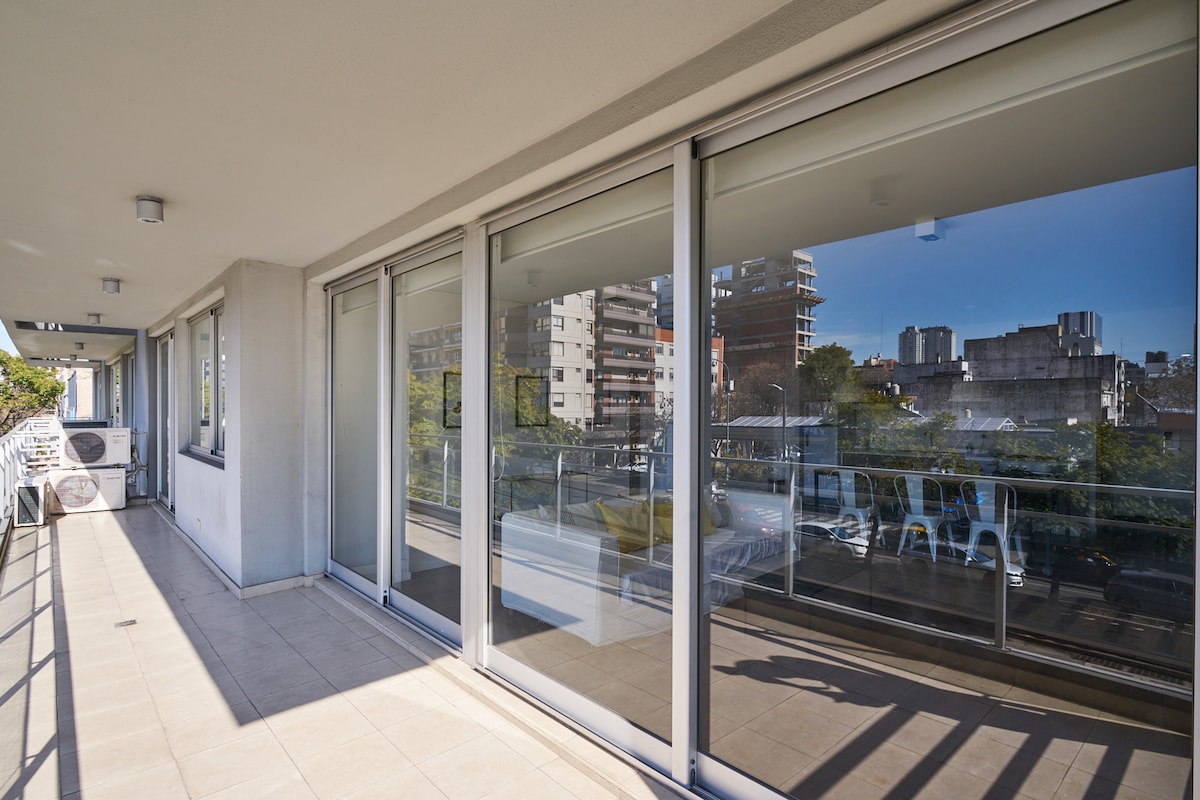 A spacious balcony is showcased, featuring large glass doors that allow ample natural light. The tiled floor enhances the area, while outside views of greenery and surrounding buildings are visible. Furniture arrangements in the background suggest a comfortable outdoor seating area.
