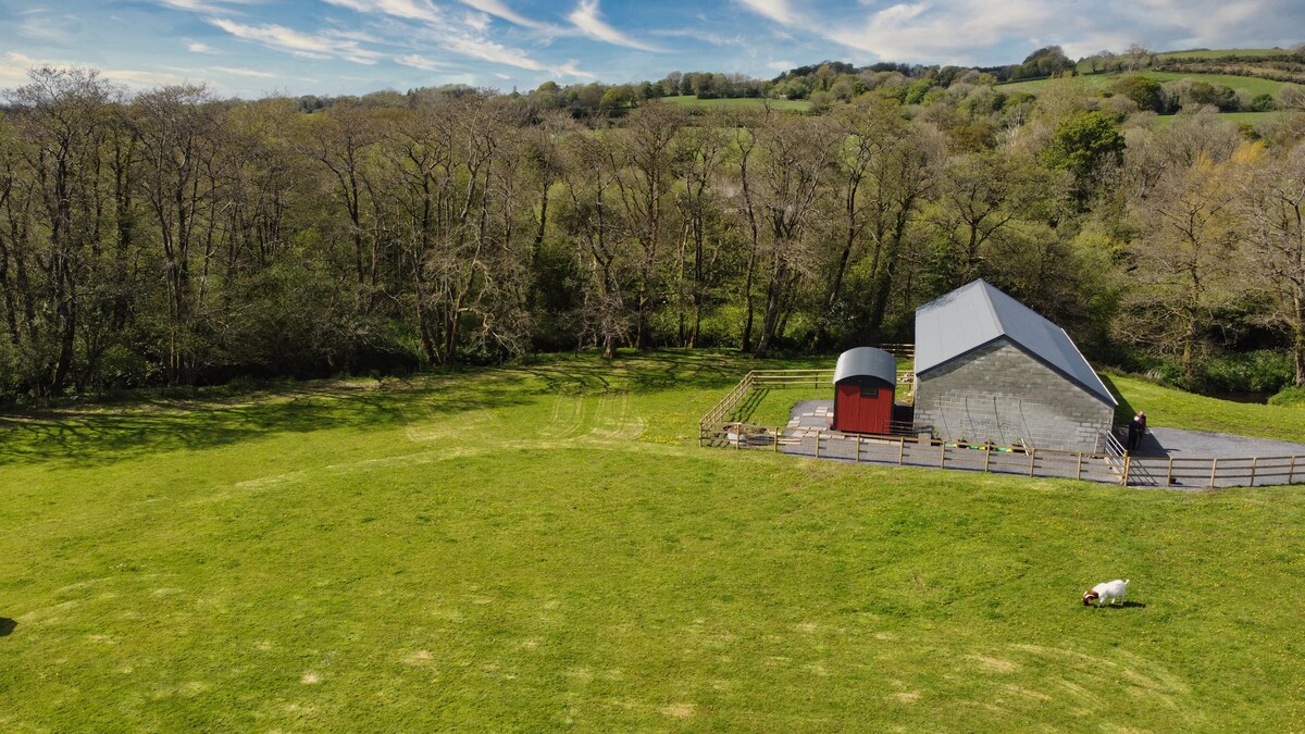 An aerial view captures a rural property surrounded by lush greenery. The modern building features a distinct roofline and is positioned next to a wooden fence. Open fields and tree lines create a serene atmosphere, with hints of nearby gentle slopes in the background.