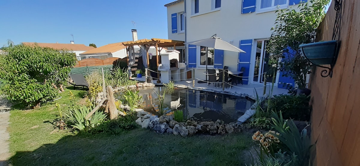 A well-maintained garden features a small pond surrounded by assorted plants and flowers. A wooden pergola offers shaded seating, while a patio leads to the light-colored house adorned with blue shutters. A clear sky complements the tranquil outdoor setting.