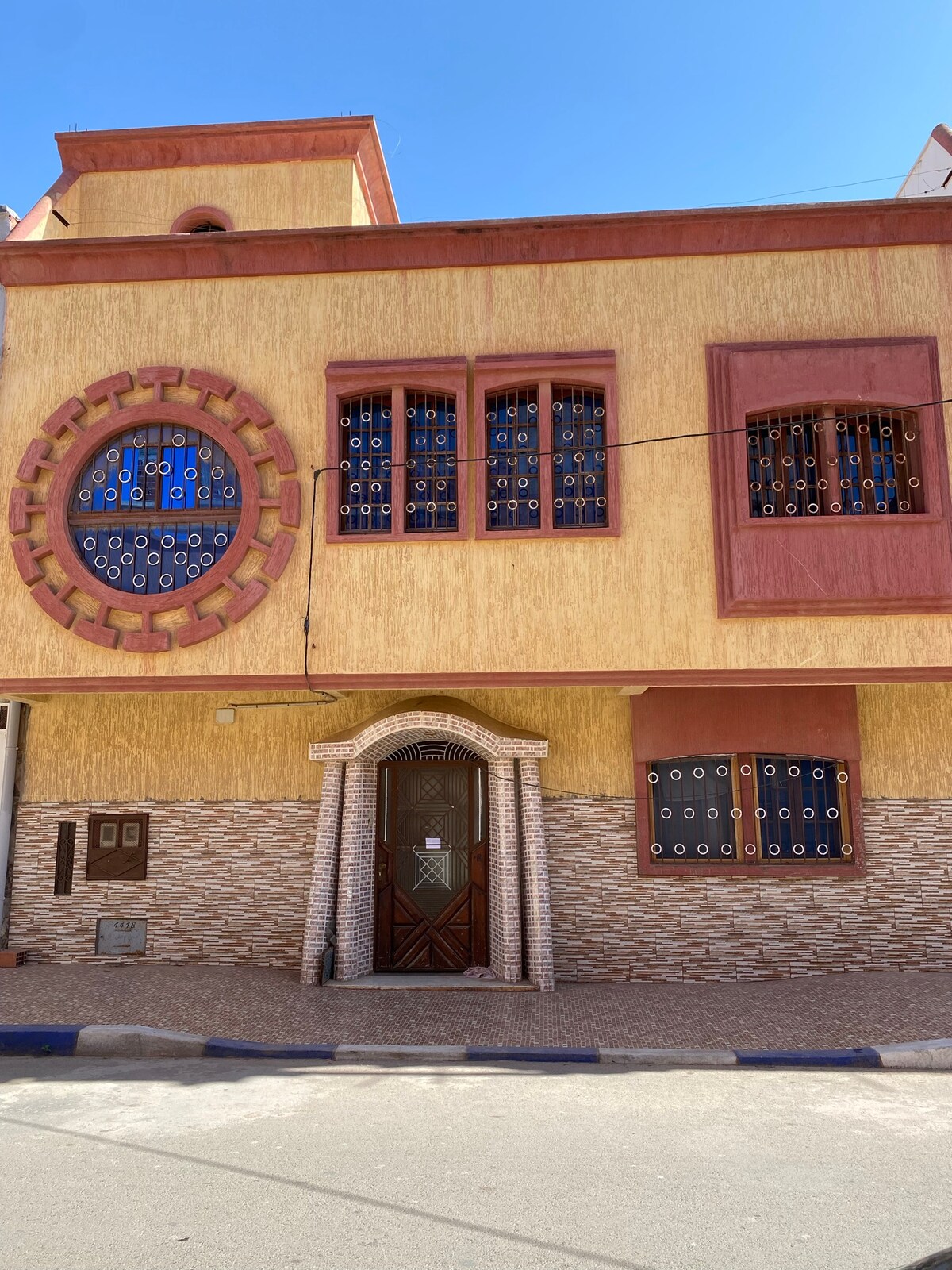 The exterior of a two-story building is displayed, featuring a vibrant yellow façade with decorative motifs. Multiple windows with intricate metal grilles provide character, and a wide wooden door is centered at the entrance. The ground is lined with patterned tiles.