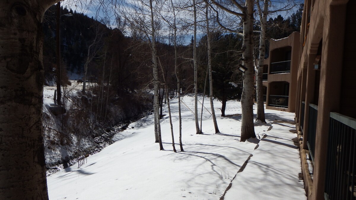 A snow-covered landscape is visible, with a winding creek bordered by bare trees. The ground is blanketed in white, while the surrounding hills reveal patches of greenery under a clear blue sky. The condo building is visible to the right, showcasing a harmonious blend with nature.
