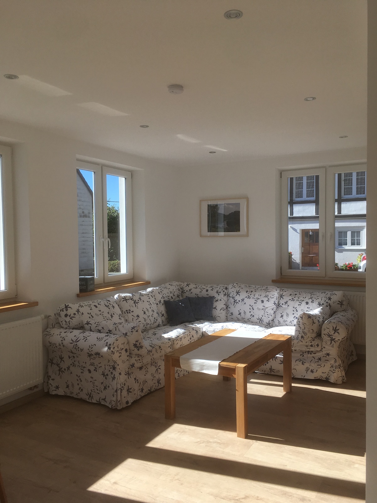 A bright living area features a corner sofa adorned with floral patterns and a wooden table. Natural light floods in through two large windows, enhancing the airy ambiance. A framed picture hangs on the wall, adding a touch of decor to the neutral space.