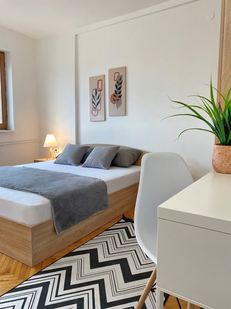A bedroom features a spacious bed with soft gray linens and decorative pillows. Artwork is displayed on the wall, and a bedside lamp provides gentle lighting. A small desk and chair are positioned beside the bed, complemented by a decorative plant on the desk.