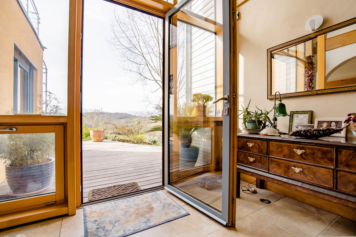 A welcoming entryway is visible, featuring a decorative wooden dresser and a mirror reflecting natural light. An open door leads to a wooden deck where greenery is visible. Potted plants are placed near the entrance, creating a connection with the outdoor environment.