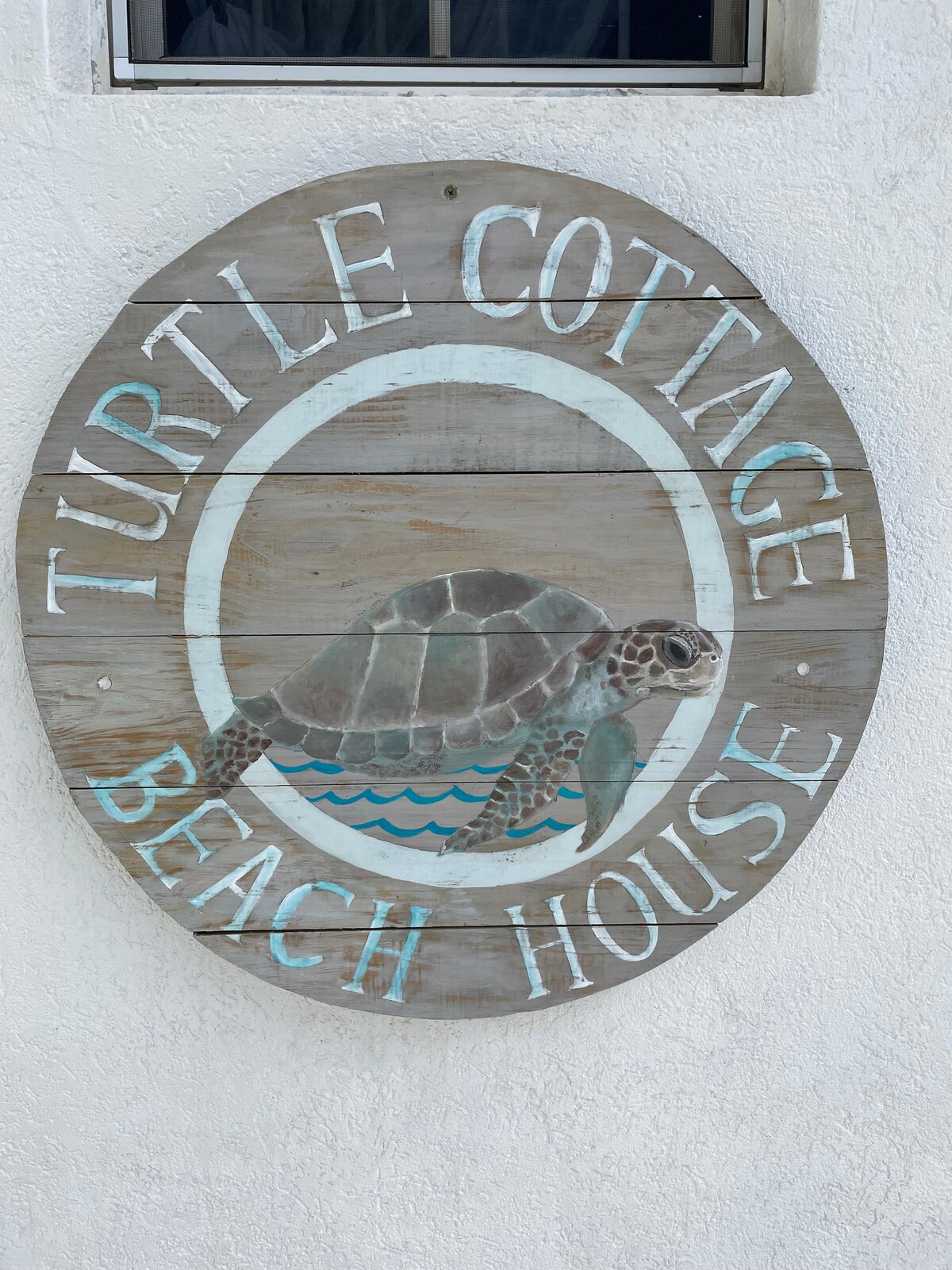 A round, wooden sign features the text 'Turtle Cottage Beach House' around an illustration of a sea turtle swimming over stylized waves. The light-colored sign contrasts with the white wall, adding a friendly entrance point to the beach cottage.