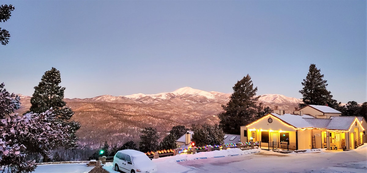 A snow-covered landscape reveals a warm yellow-lit building nestled against the backdrop of Sierra Blanca Peak. The surrounding trees, adorned with a dusting of snow, enhance the serene mountain setting, while the twilight sky casts a soft glow over the entire scene.