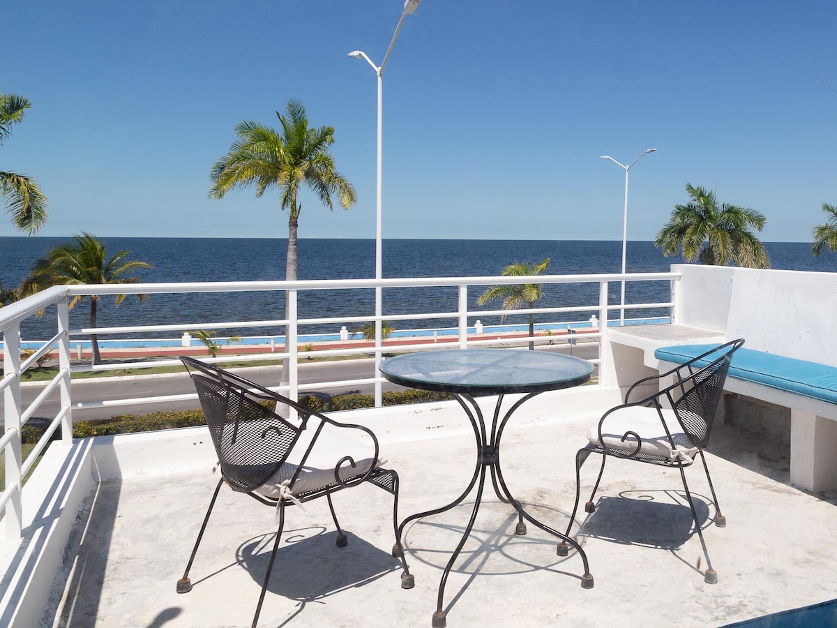 A rooftop terrace features a round table surrounded by two black chairs, overlooking a vast sea. Palm trees sway gently in the foreground, and a clear blue sky stretches above, creating a serene atmosphere for relaxation.