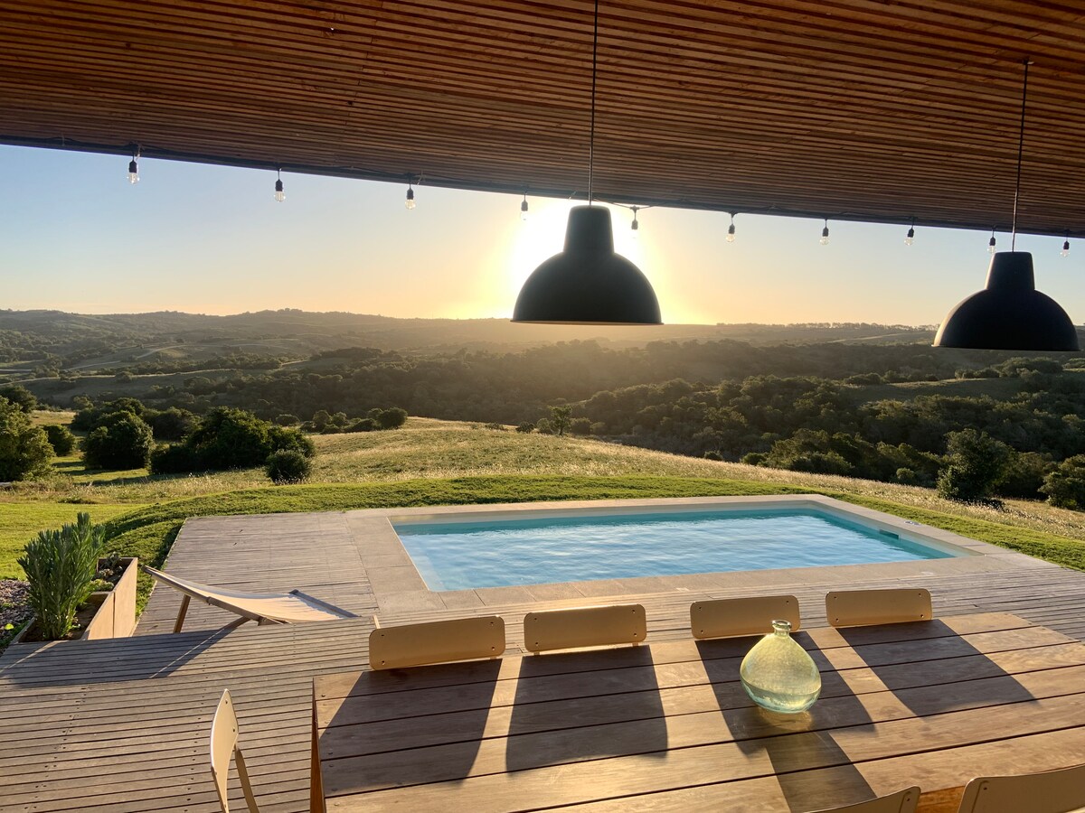 A private swimming pool is framed by a wooden deck, with seating arranged around a central glass vase. Overhead, pendant lights hang from a wooden ceiling. The expansive view of rolling hills under a warm sunset is captured in the background.