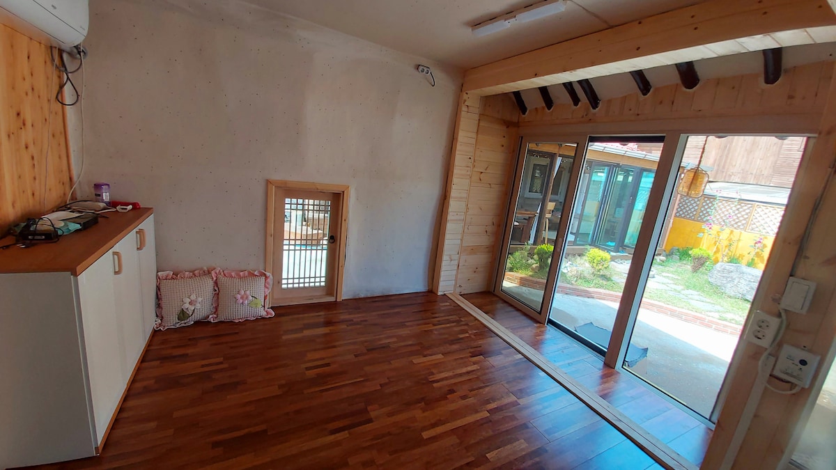 A light-filled interior features wooden flooring and a neutral color palette. Large glass doors open to the outdoors, allowing natural light to illuminate the space. A small window and wall-mounted air conditioning unit are also visible, along with a practical storage cabinet.