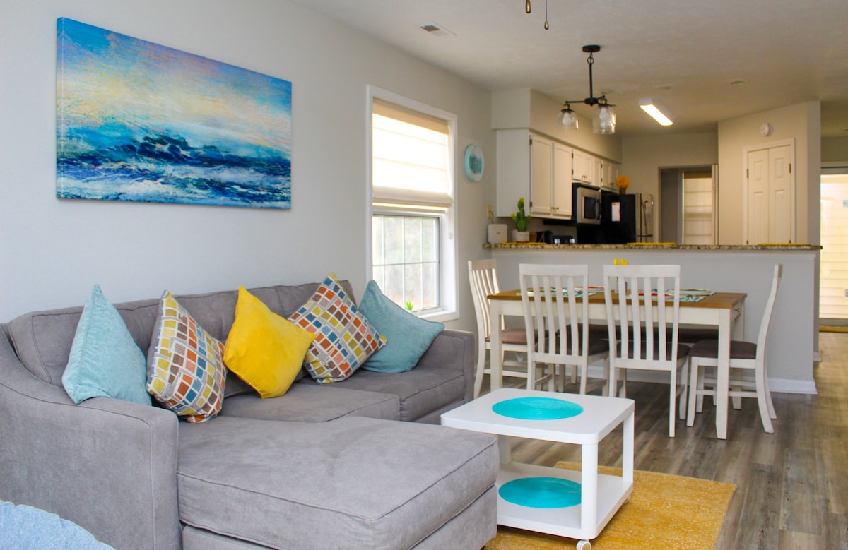 A spacious living area is furnished with a large sectional sofa adorned with colorful throw pillows. A coffee table is positioned in front, and a dining table with white chairs is set in the background. Sunlight enters through nearby windows, illuminating the modern decor.