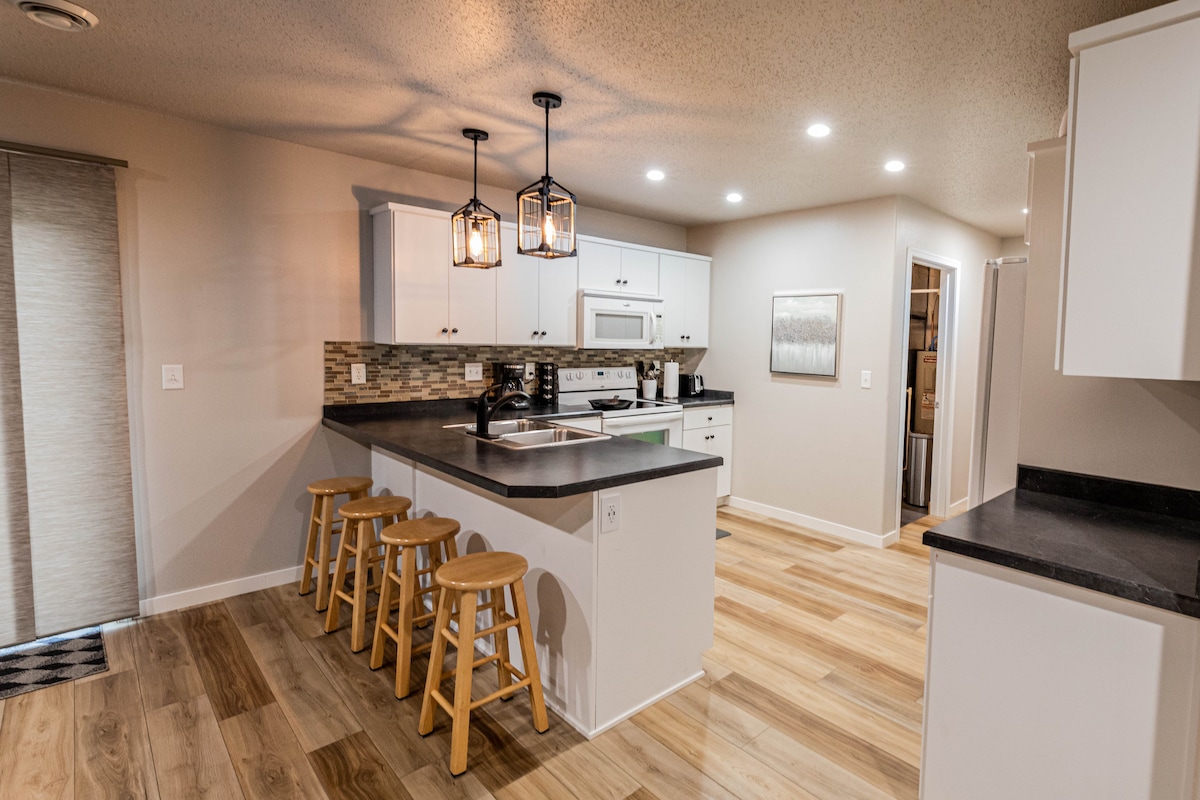 The kitchen area features a modern design with white cabinetry and dark countertops. Three wooden stools are positioned at the kitchen island, which provides ample workspace. Soft lighting from hanging fixtures illuminates the room, highlighting the combination of natural wood flooring and tiled backsplash.