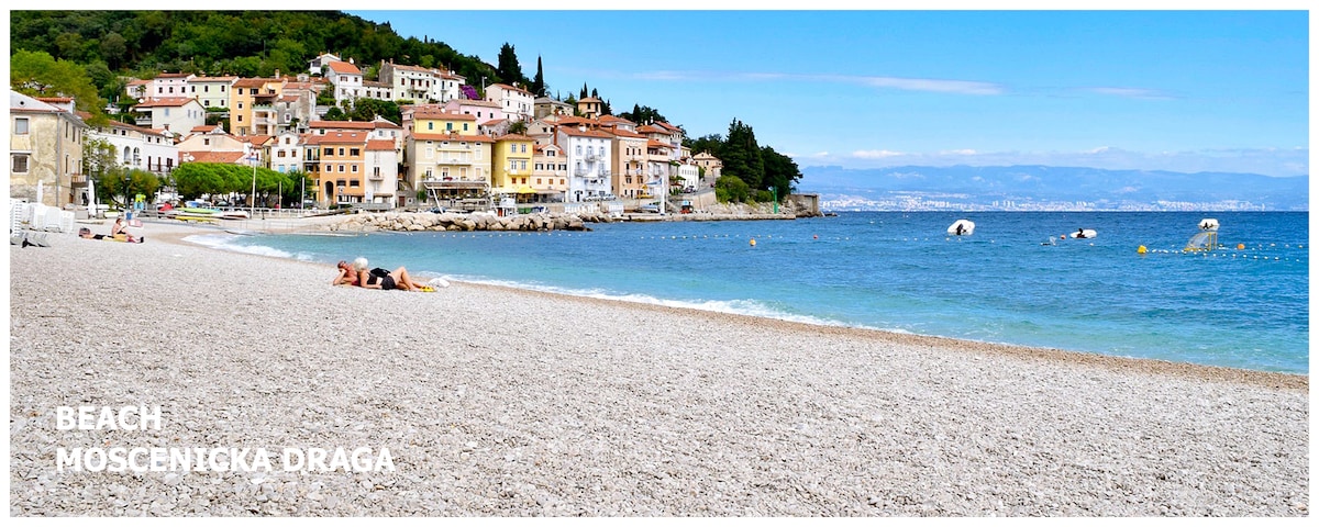 A serene beach scene features a pebbled shoreline leading to calm blue waters. Colorful buildings line the coast, set against a backdrop of green hills. Two individuals relax on the beach, enjoying the sun and the peaceful atmosphere.