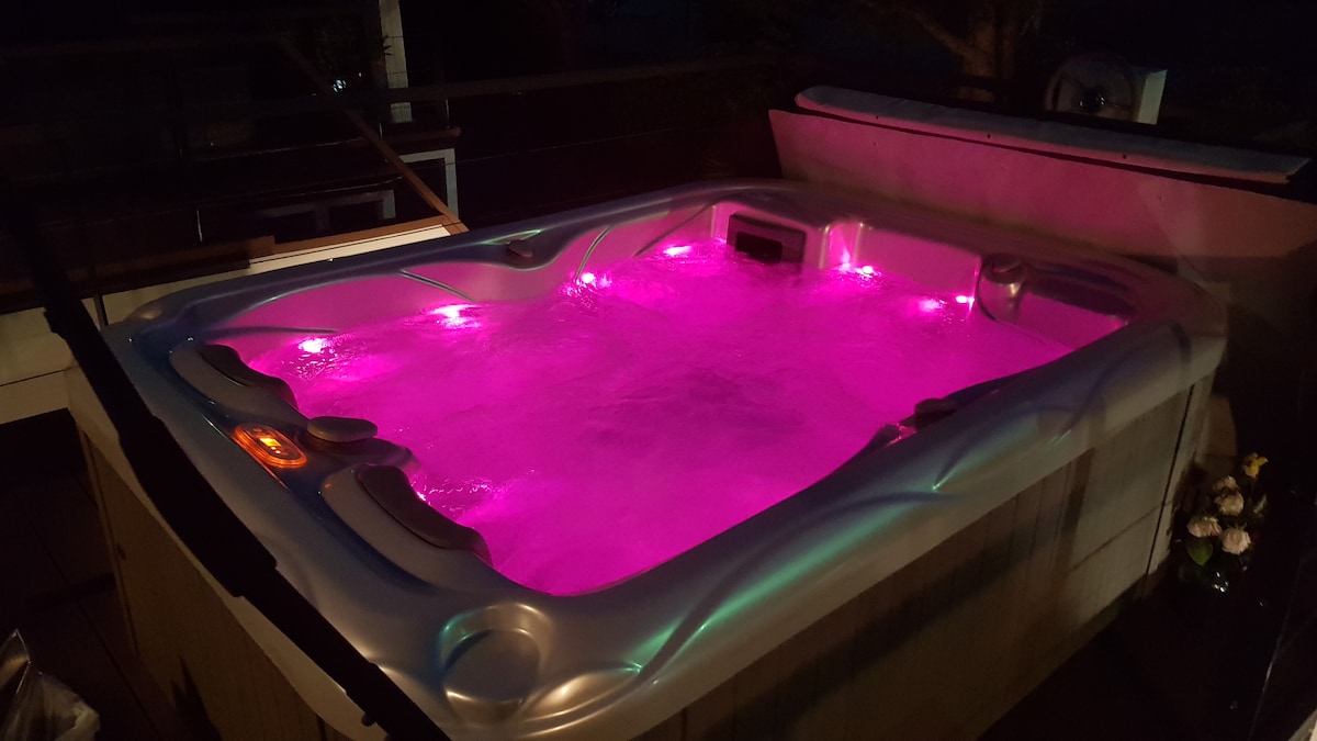 A private hot tub is featured on the terrace, illuminated with soft pink and blue lights. The modern design offers comfortable seating with jets, set against a backdrop of a tranquil outdoor space. The ambiance invites relaxation and enjoyment.