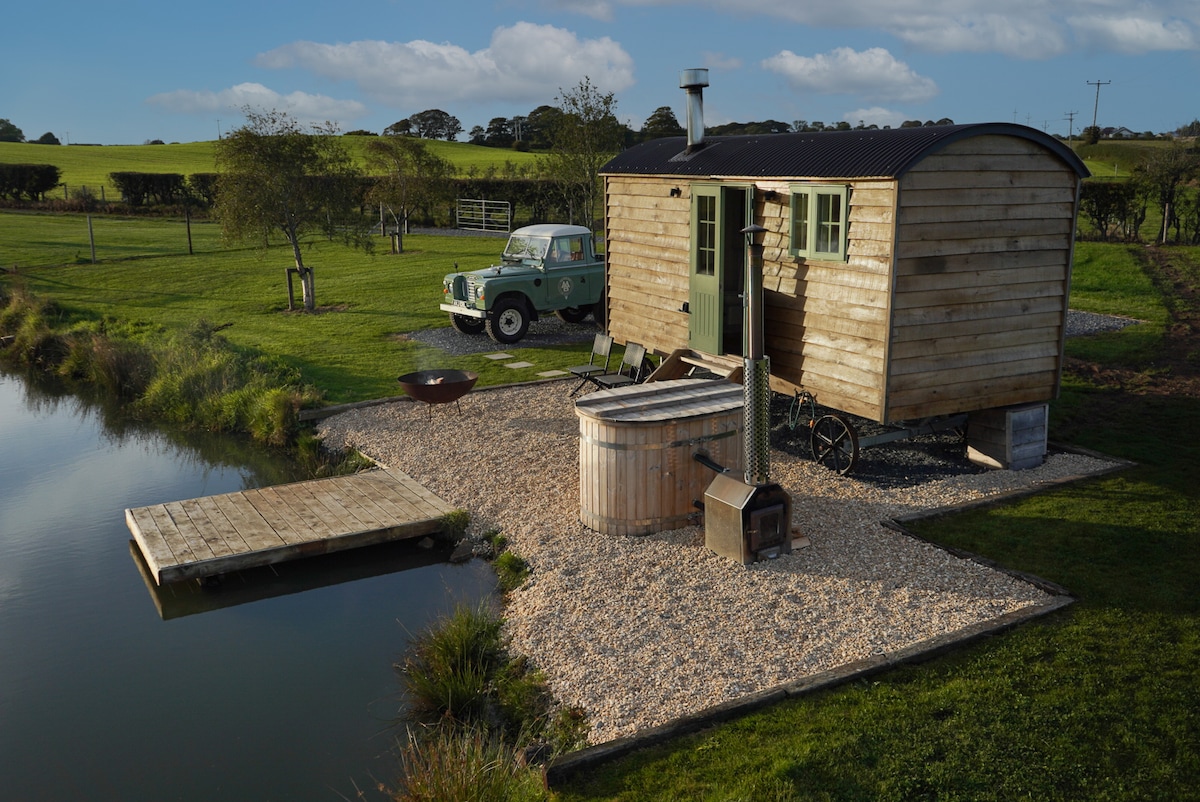 A luxury shepherd's hut is positioned near a private lake, featuring a wood-fired hot tub and a small wooden dock. An vintage green vehicle is parked nearby, amidst expansive grassy surroundings and trees, creating a serene rural setting.