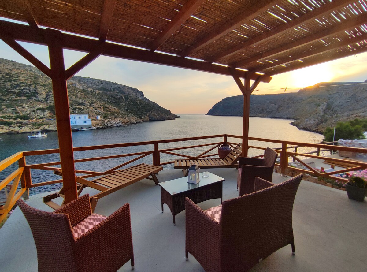 A spacious terrace with wooden beams provides a relaxing sitting area, featuring comfortable wicker chairs and a low table. The view encompasses calm waters and rocky cliffs, with a sunset creating a warm glow over the landscape.