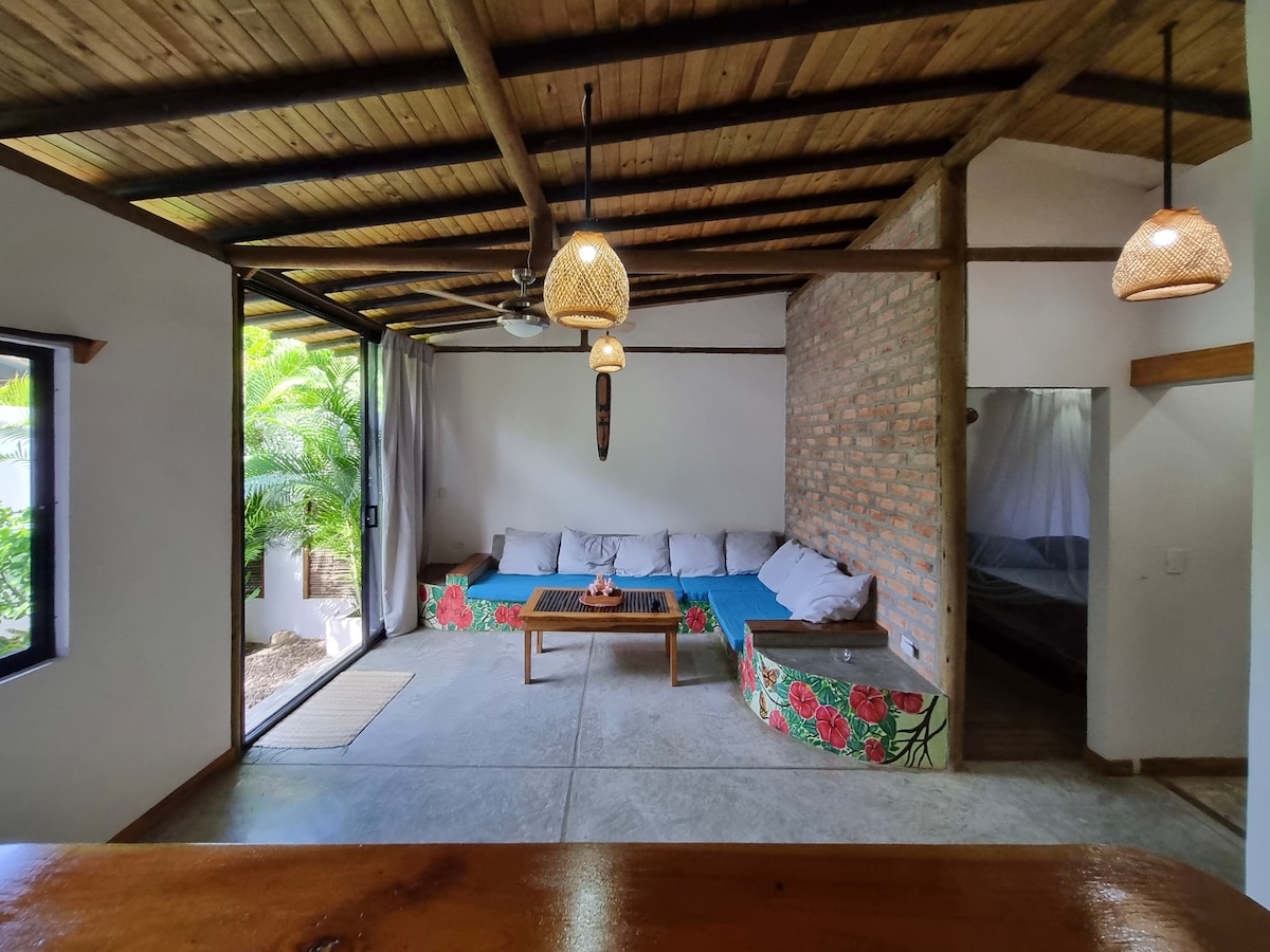 A spacious living area features wooden beams across the ceiling and large windows allowing natural light. A comfortable seating arrangement is adorned with colorful floral patterns, providing a cheerful contrast against the minimalist interior. Nearby, an entrance to a bedroom can be seen, suggesting easy access.