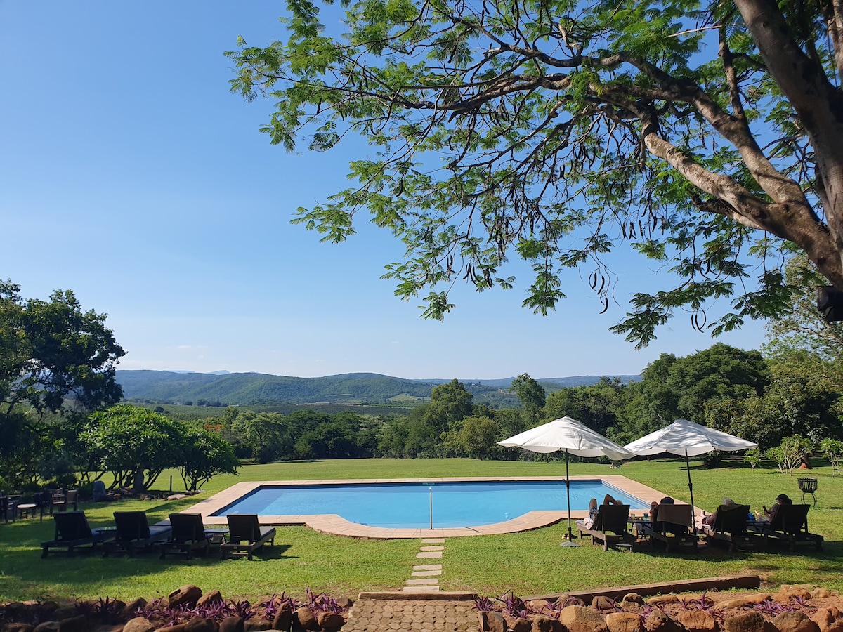 A serene outdoor area features a large swimming pool surrounded by well-maintained grass. Sun loungers are positioned nearby under shaded umbrellas, with a panoramic view of rolling hills and trees in the background, creating a peaceful environment.