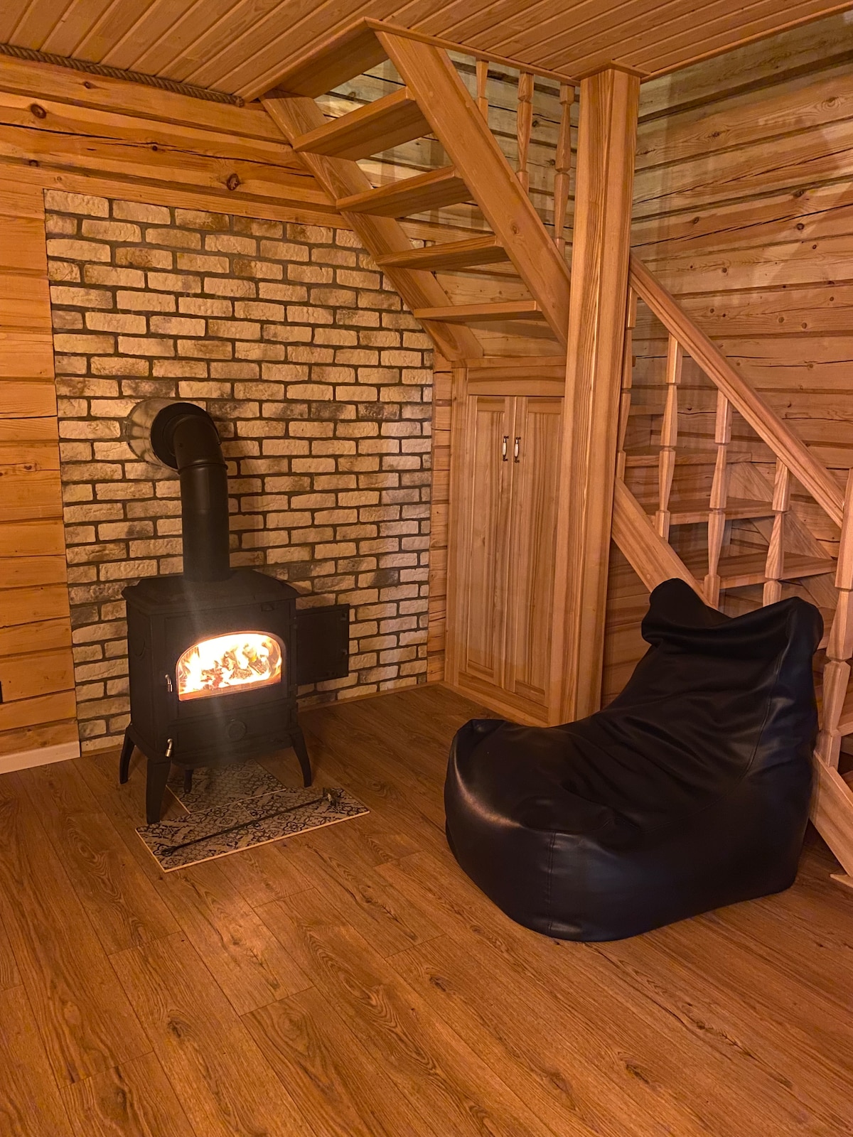 A warm and inviting interior features a wood-burning stove with a black chimney against a brick wall. Natural wood accents are present throughout, complemented by a cozy black bean bag chair and a staircase leading to the upper level. Wooden flooring adds to the rustic atmosphere.