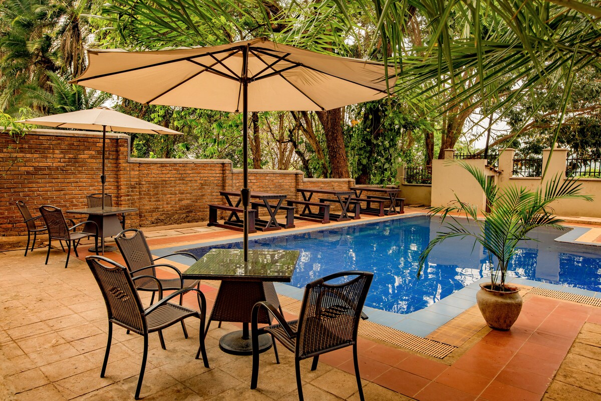 An outdoor area features a serene swimming pool surrounded by greenery. Comfortable black chairs and tables are positioned under large umbrellas, providing shaded seating. A decorative plant is situated near the poolside, enhancing the inviting atmosphere of this relaxing space.