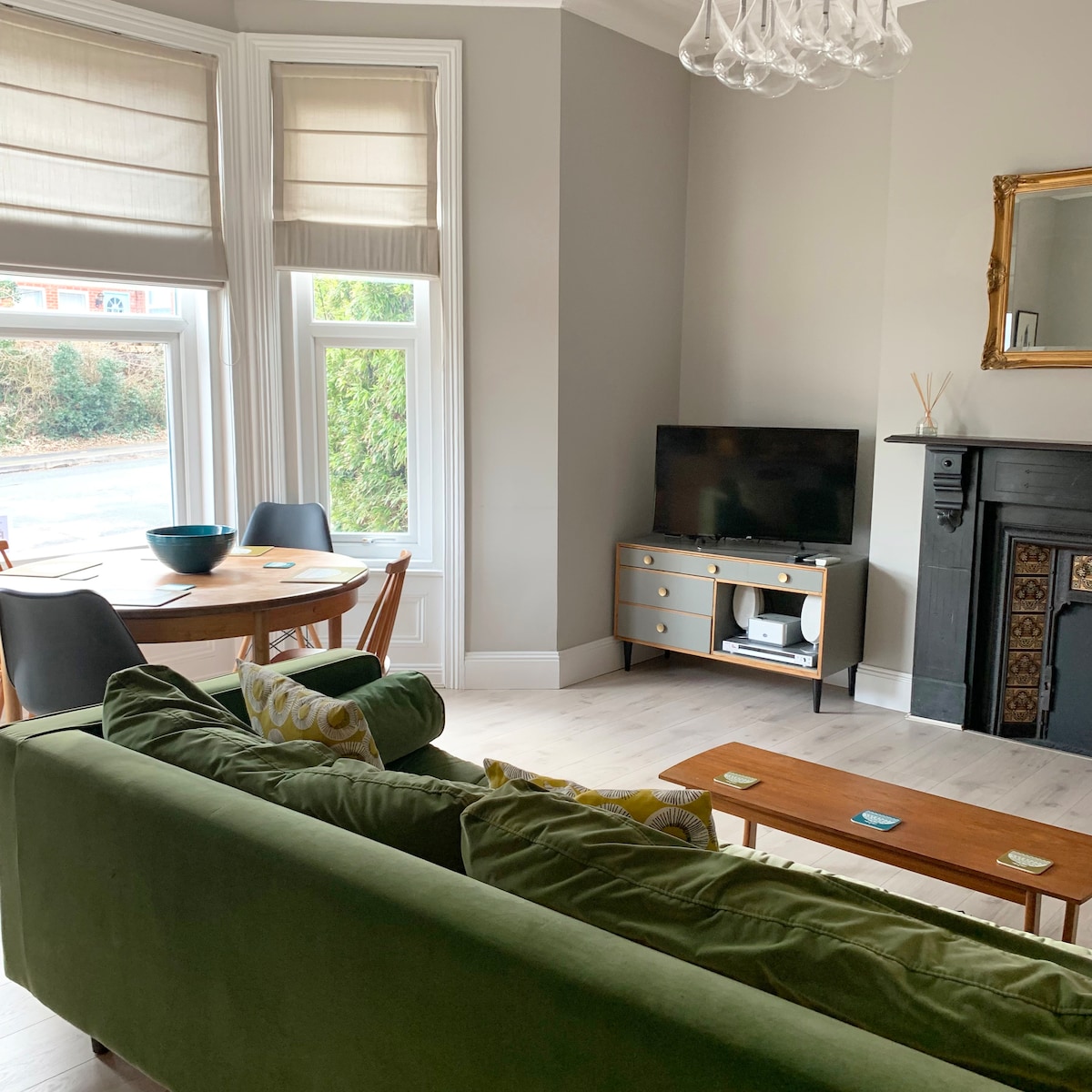 A spacious living area features a green sofa facing a low coffee table and a modern flat-screen TV. A round dining table with chairs is positioned near large windows, allowing natural light to fill the room. Wall decor includes a decorative mirror above a fireplace.