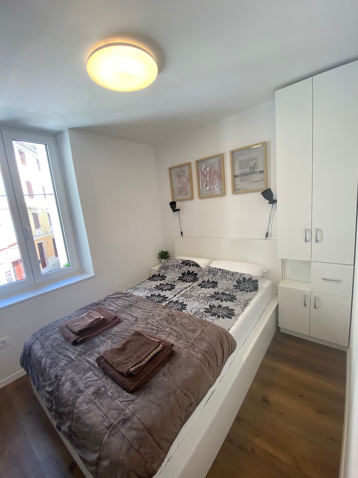 A bedroom is presented with a queen bed, dressed in a floral patterned coverlet. Two towels are neatly placed on the bed. Nightstands with reading lights are positioned on either side. A window allows natural light to fill the space, framed by light walls and wooden flooring.