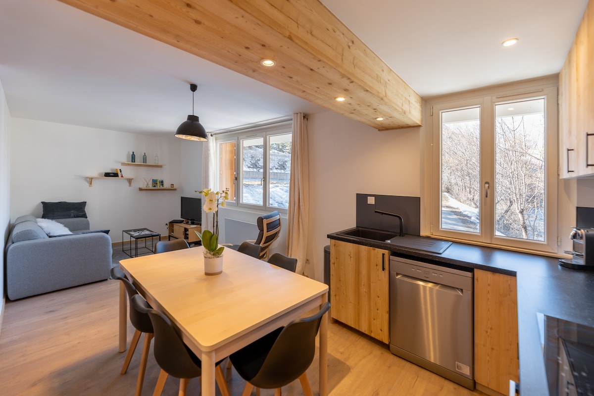 A bright and modern living space is shown, featuring a cozy couch and a wooden dining table surrounded by black chairs. Natural light pours through large windows, revealing a picturesque outdoors. The kitchen area includes an equipped counter with a dishwasher and various appliances.