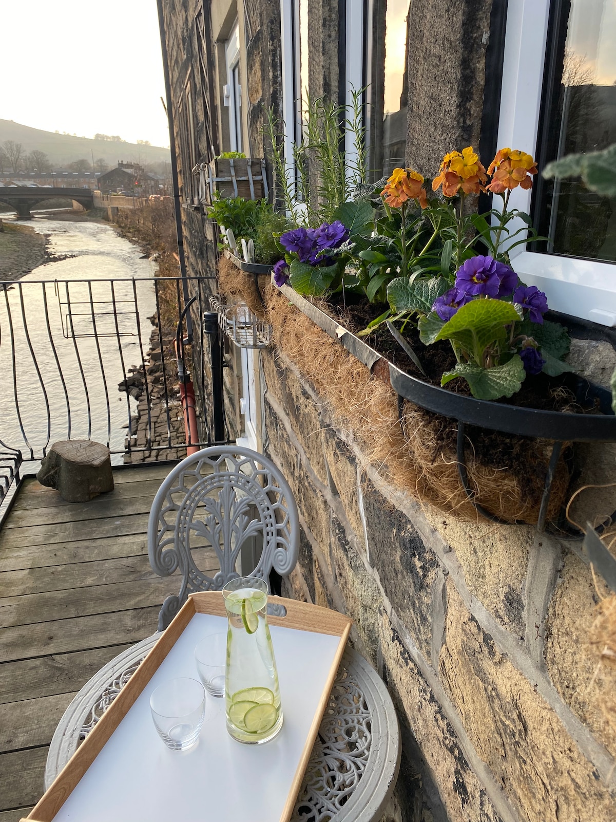 A small balcony features a vintage-style table and chair set, accompanied by a decorative tray with a glass and a bottle. Colorful flowers bloom in window boxes, adding a touch of nature to the wooden deck overlooking the River Calder.