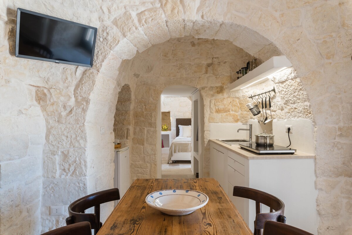 An arched entryway leads into a cozy dining area featuring a wooden table surrounded by dark chairs. A stone wall adds rustic charm, illuminated by soft lighting. A retro television is mounted on the opposite wall, while modern kitchen amenities are visible in the open layout.