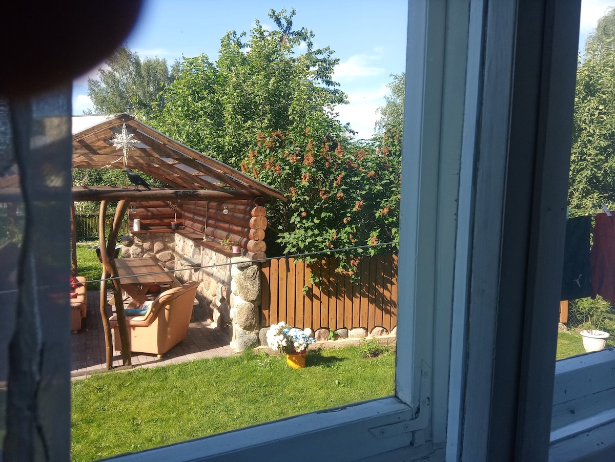 A view from a window showcases a tranquil outdoor area featuring a wooden gazebo and a dining table surrounded by greenery. Bright flower pots and a clothesline are visible, creating a serene garden setting. The landscape is enhanced by the clear sky above.