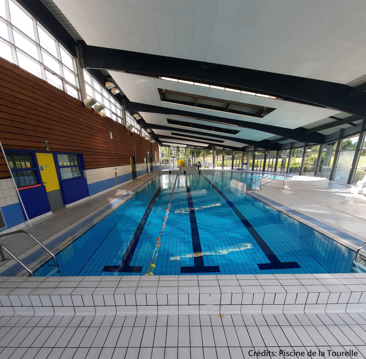 An indoor swimming pool is featured, showcasing clear blue water surrounded by tiled flooring. Large windows allow natural light to fill the space, with a seating area visible on the side. The pool is lined with lane markers and diving blocks.