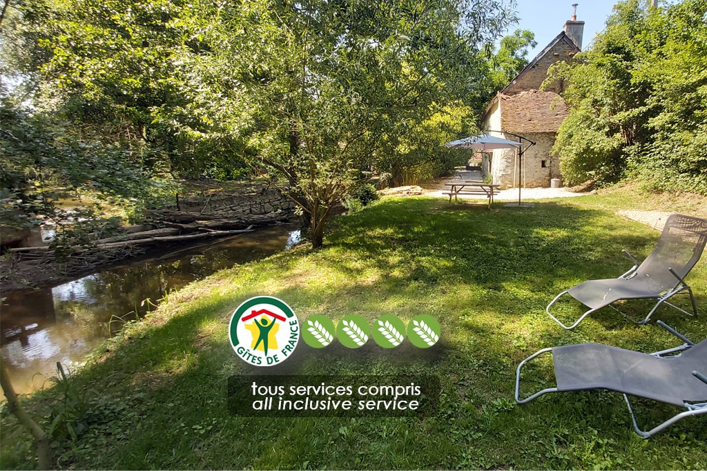 A serene outdoor space features a grassy area beside a tranquil river. A picnic table with an umbrella provides shaded seating, while lounge chairs are positioned nearby. The rustic house blends harmoniously with the surrounding greenery, creating a peaceful retreat.