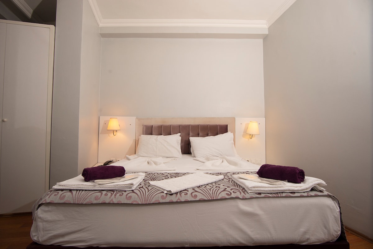 A spacious bed is centrally positioned against a soft gray wall, featuring plush pillows and folded towels. Two bedside lamps provide gentle lighting, enhancing the room's calming ambiance. Minimalistic furniture complements the inviting layout, ideal for relaxation.