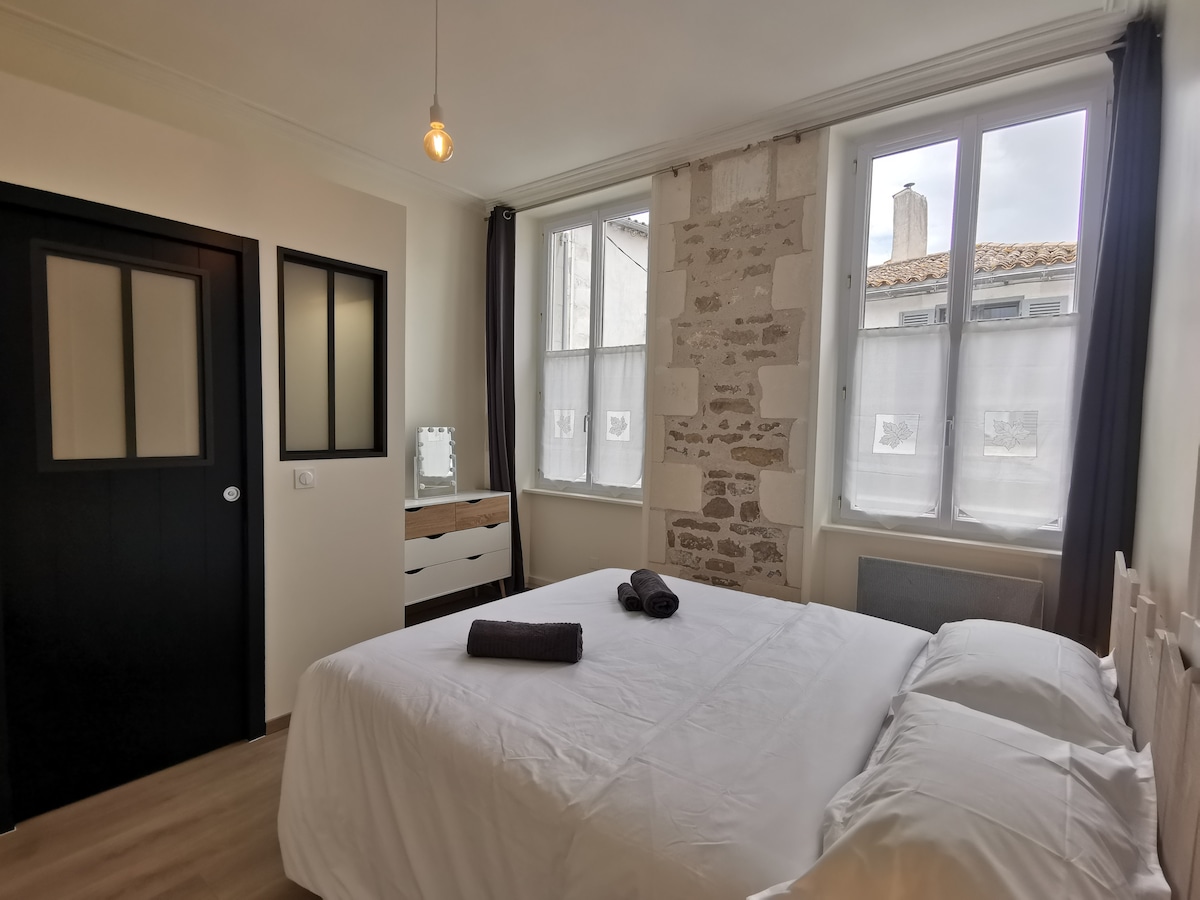 A spacious bedroom features a large bed with crisp white linens. Natural light floods the room through tall windows, which are dressed in light, sheer curtains. A mirrored dresser is positioned against a textured stone wall, and two dark towels are neatly placed on the bed.