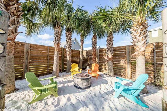 A secluded outdoor area is surrounded by palm trees and wooden fencing. Four colorful adirondack chairs are arranged around a stone fire pit, with soft white sand covering the ground, creating a comfortable space for relaxation and gatherings.