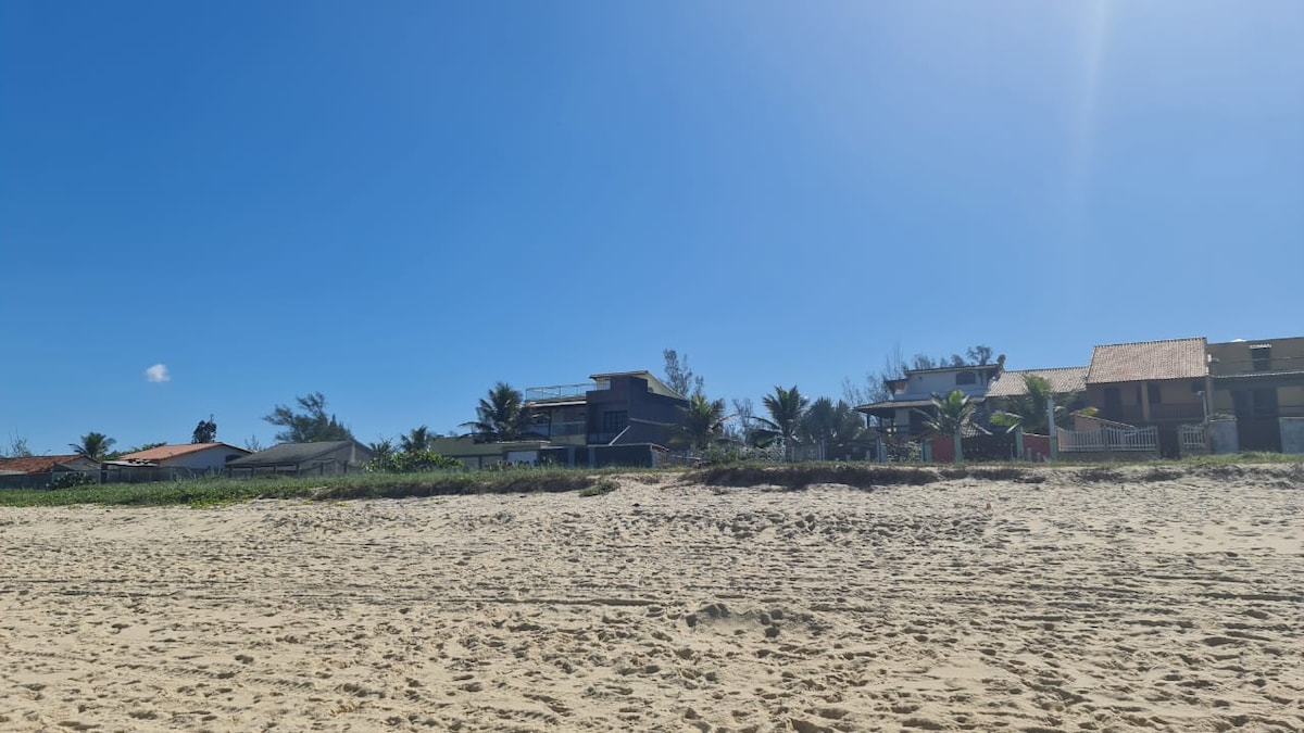 A clear blue sky is visible above a sandy beach, where several homes with tropical landscaping are lined beside the shore. Palm trees and green vegetation are seen in the background, contributing to the serene beach environment.