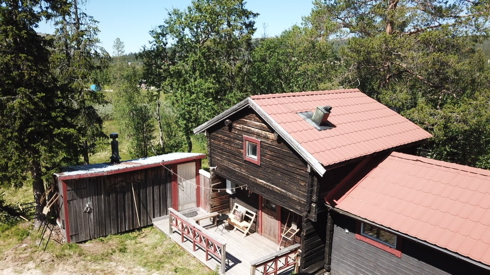 An aerial view captures a charming wooden cabin with a red tile roof, surrounded by lush greenery. The deck features seating for relaxation, while a storage shed is visible on the side. The natural landscape creates a serene backdrop.