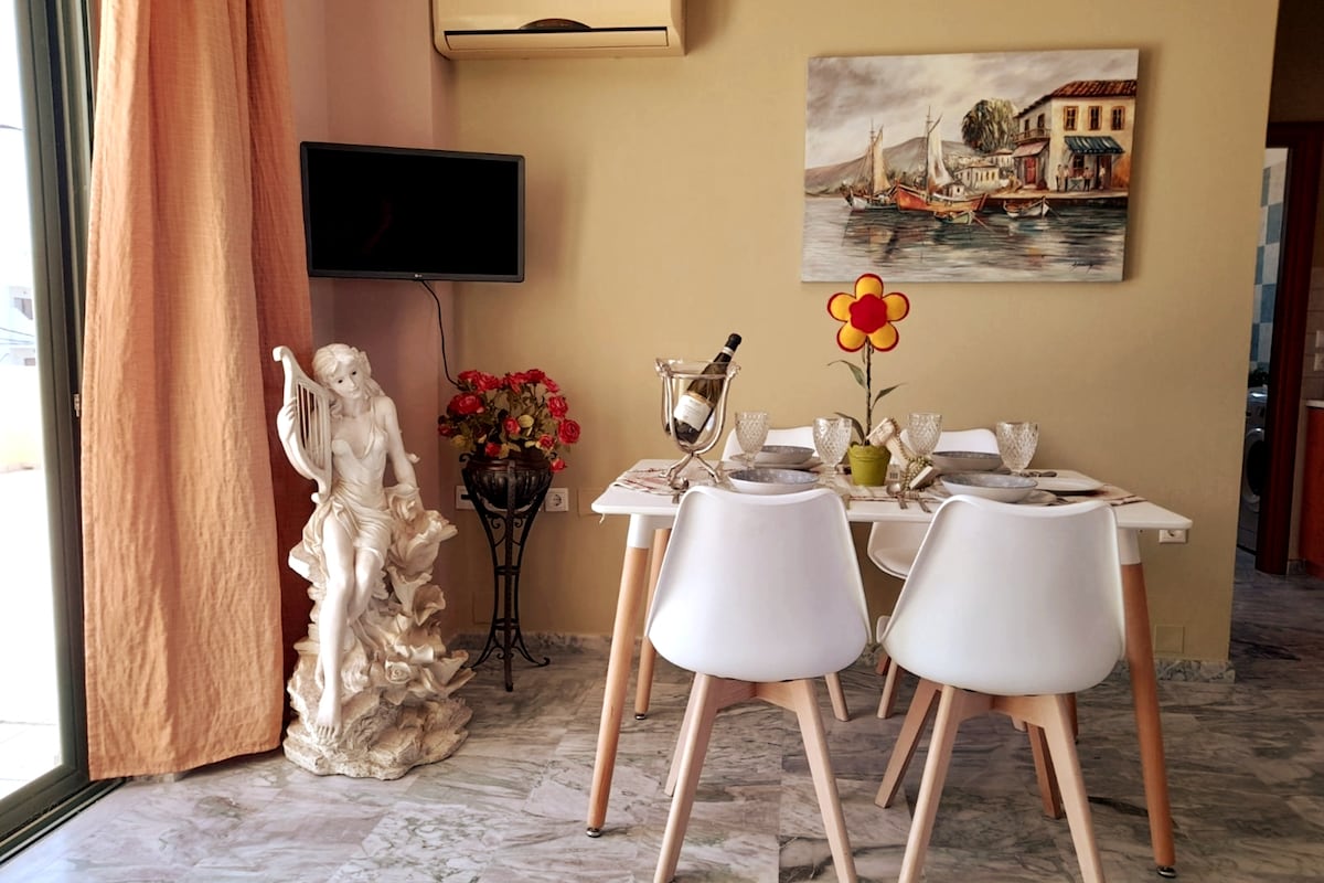 A dining area is presented with a modern white table set for four, featuring elegant dishes and a flower centerpiece. A wall-mounted television is visible. A decorative sculpture adds character to the space, while soft curtains frame a window, allowing natural light to enhance the atmosphere.